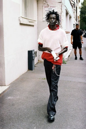Model walking outdoors along a street wearing a layered cream and red knit sweater paired with black trousers.