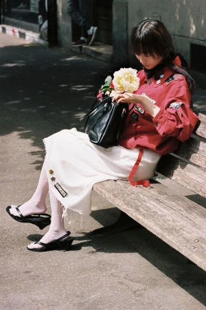 Model wearing a red jacket and white skirt sitting on a wooden park bench holding a flower and a black handbag