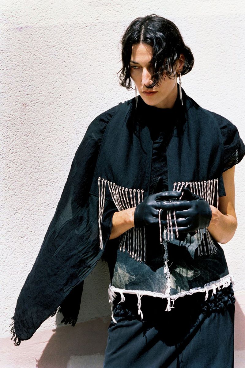 Model Alicia Lindén Guinez posing in a black chain-fringed top, black leather gloves, and draped shawl against a sunny textured wall.