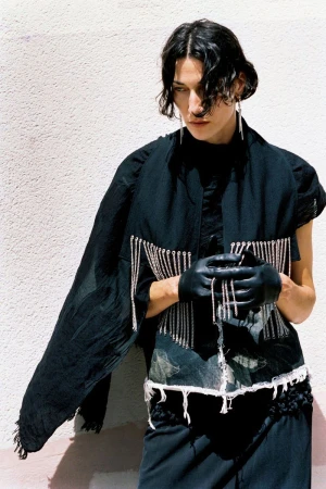 Model Alicia Lindén Guinez posing in a black chain-fringed top, black leather gloves, and draped shawl against a sunny textured wall.