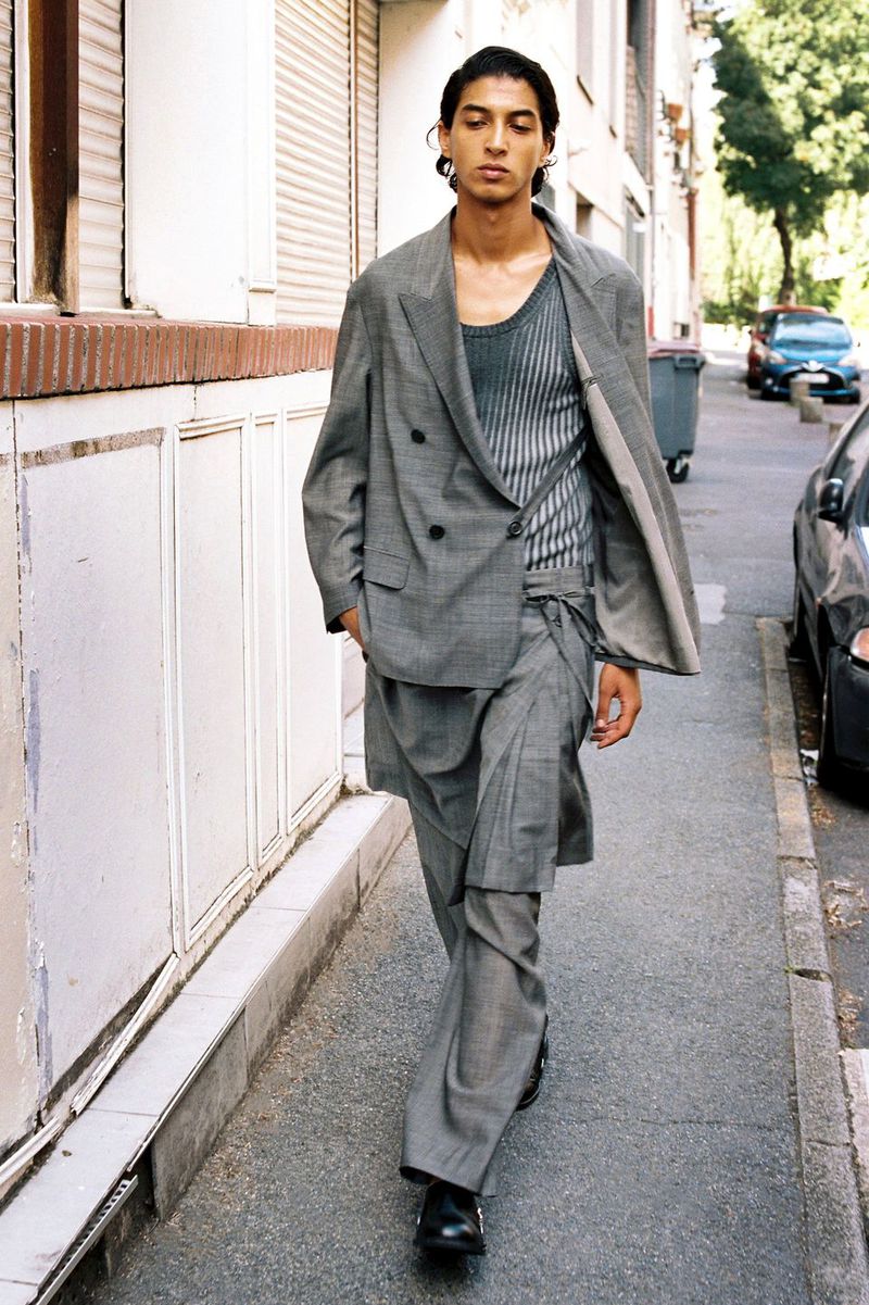Model walking down an urban sidewalk wearing a grey double-breasted blazer with layered wrap trousers and a knit scoop-neck top.
