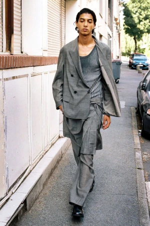 Model walking down an urban sidewalk wearing a grey double-breasted blazer with layered wrap trousers and a knit scoop-neck top.