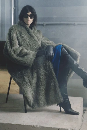 Model seated wearing a textured green faux fur coat, black sunglasses, leather gloves, and tall boots by Christian Wijnants