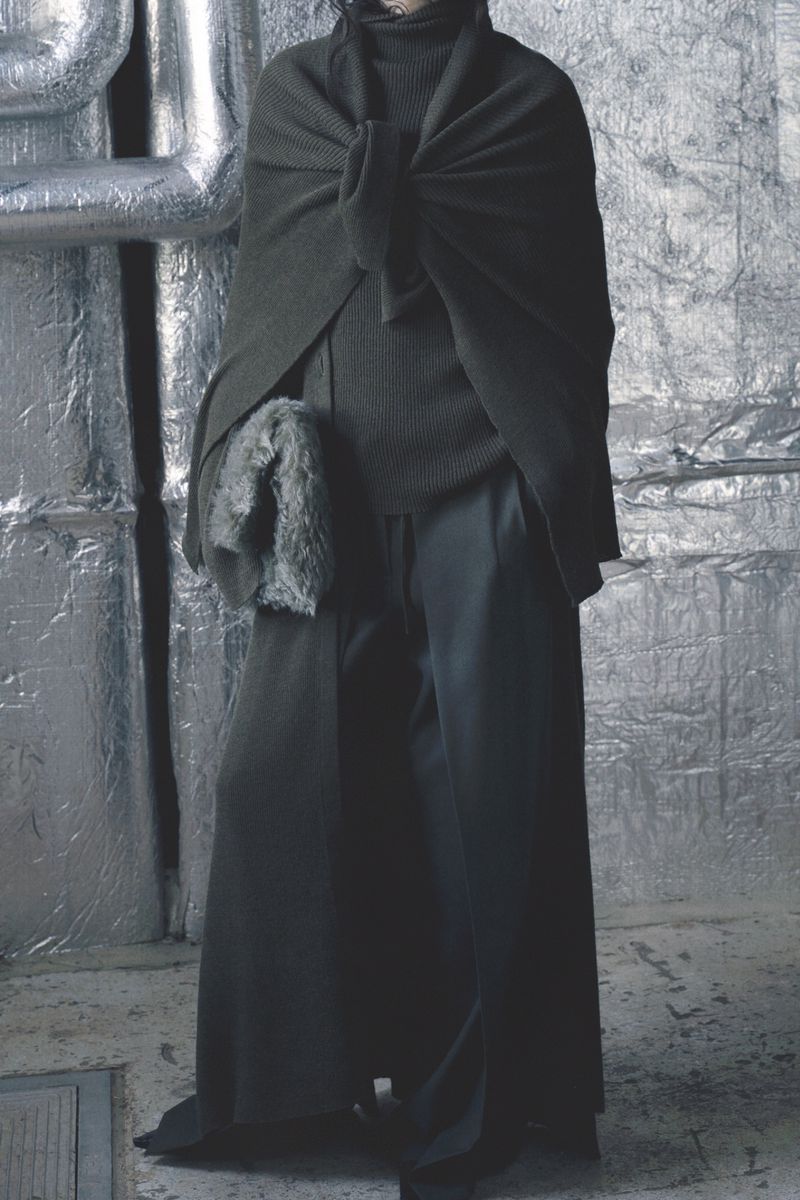 Model wearing a dark green knotted cape, ribbed turtleneck sweater, and wide-leg trousers against foil-covered insulation.