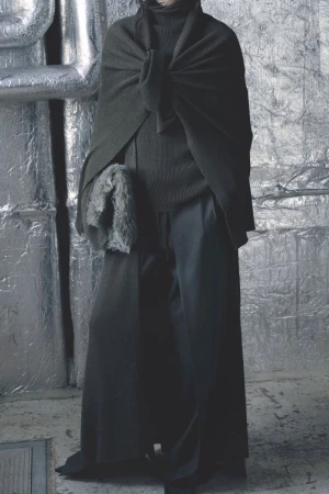 Model wearing a dark green knotted cape, ribbed turtleneck sweater, and wide-leg trousers against foil-covered insulation.