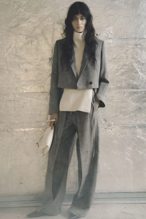 Model standing against a textured metallic background wearing a grey blazer, white turtleneck, and wide-leg trousers