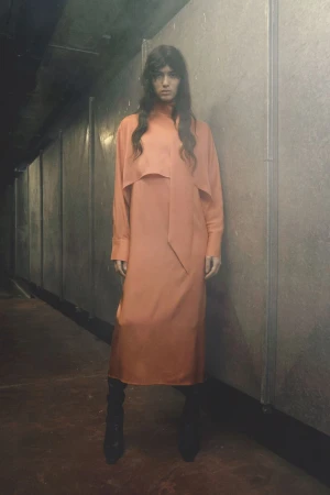 Model wearing a long orange dress standing in a corridor with metal walls for Christian Wijnants Pre-Fall 2026.