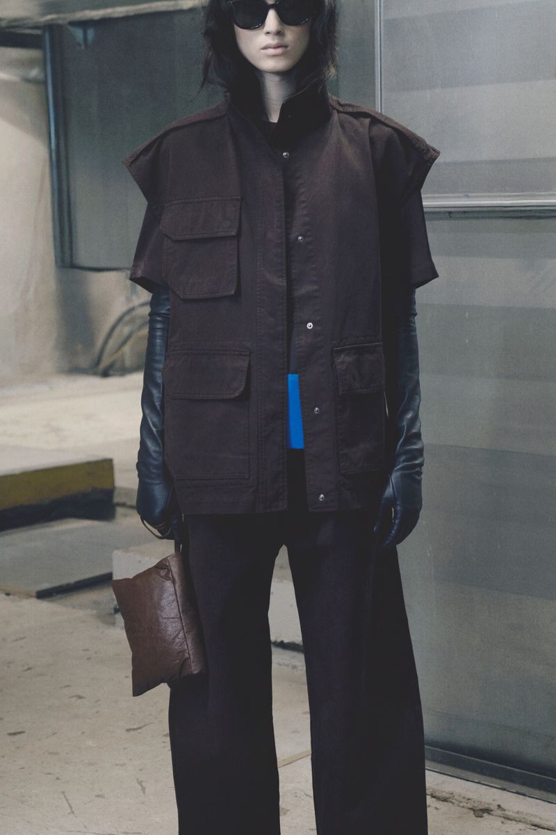 Model wearing dark sunglasses, a sleeveless brown utility jacket, long leather gloves, and dark trousers in an industrial setting.
