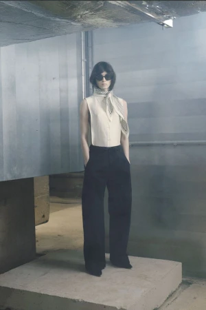 Model wearing a sleeveless cream top and black trousers with a patterned scarf standing on a concrete platform in an industrial space