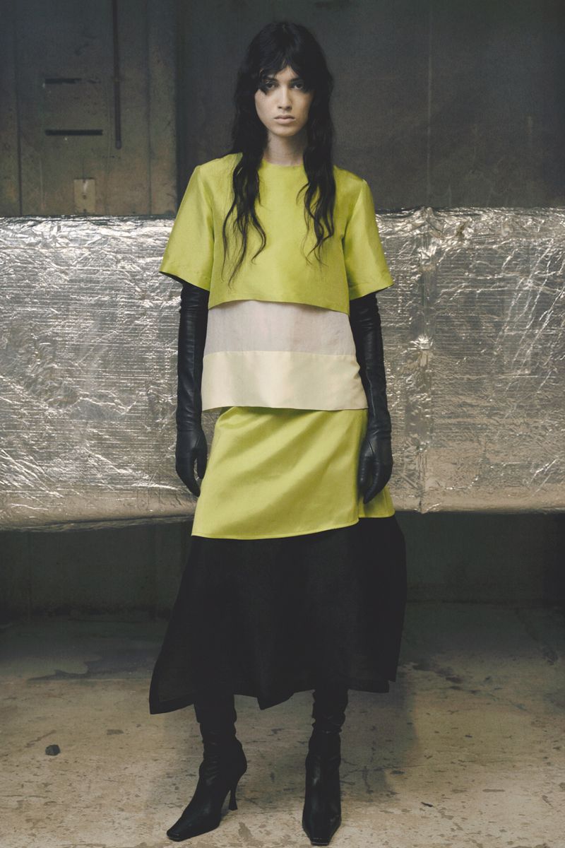 Model Svetlana Lethelier posing in a chartreuse layered top, skirt, and long black leather gloves against a foil-backed wall.