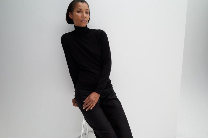 Model Romae Gordon wearing a black turtleneck sweater and black trousers seated on a stool against a white seamless studio backdrop.