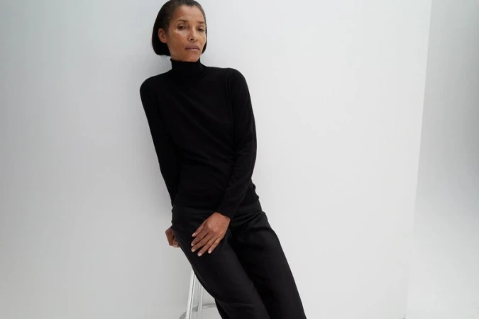 Model Romae Gordon wearing a black turtleneck sweater and black trousers seated on a stool against a white seamless studio backdrop.