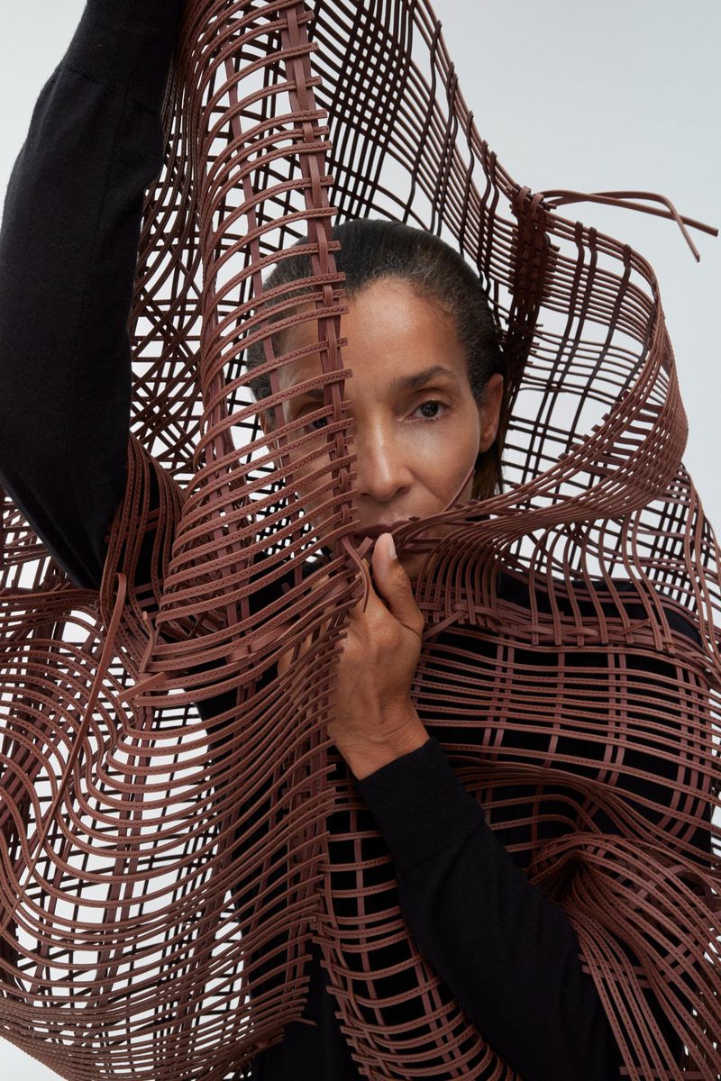 Model Romae Gordon peering through an open brown sculptural woven lattice structure wearing a black top.