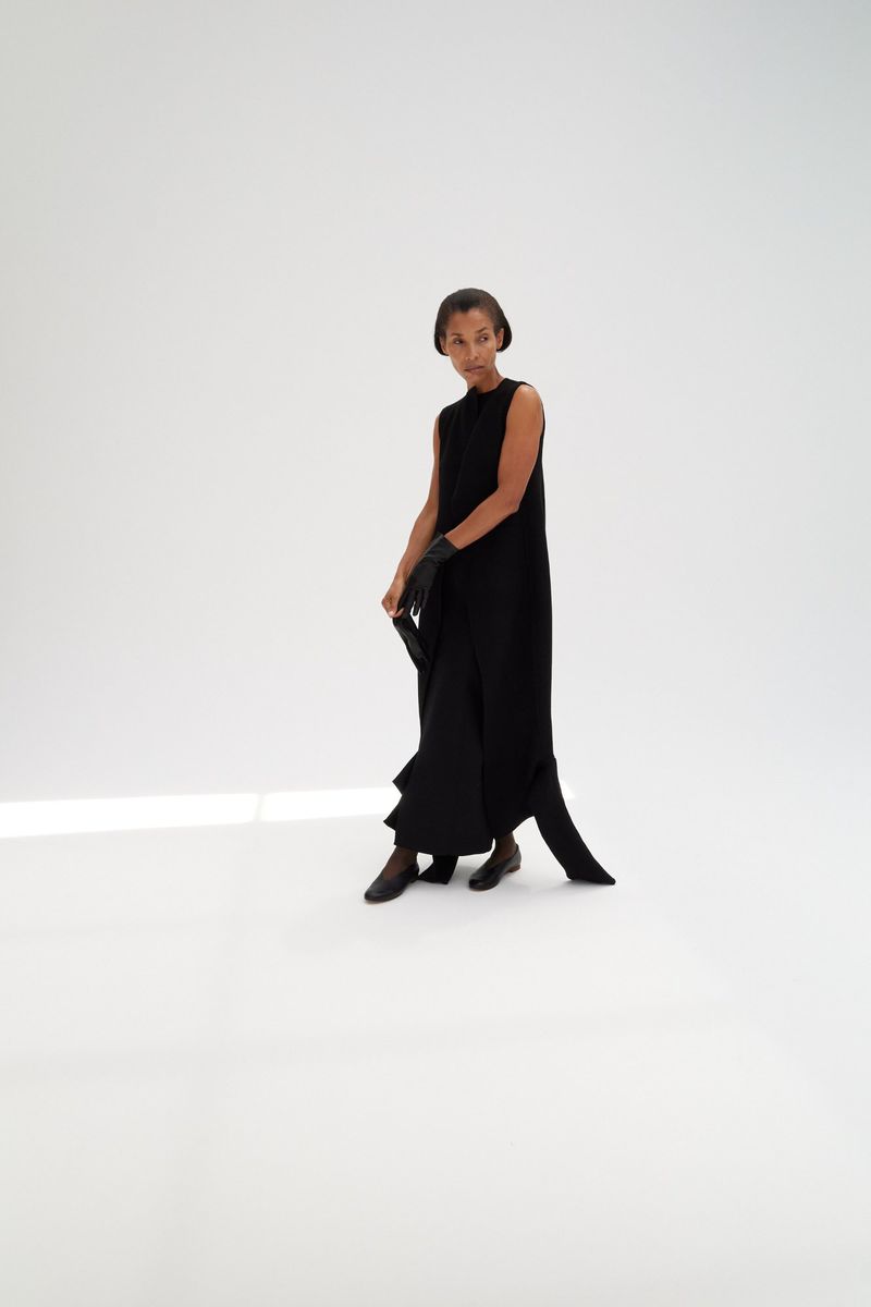 Model wearing a black sleeveless dress and leather gloves standing on a white seamless background