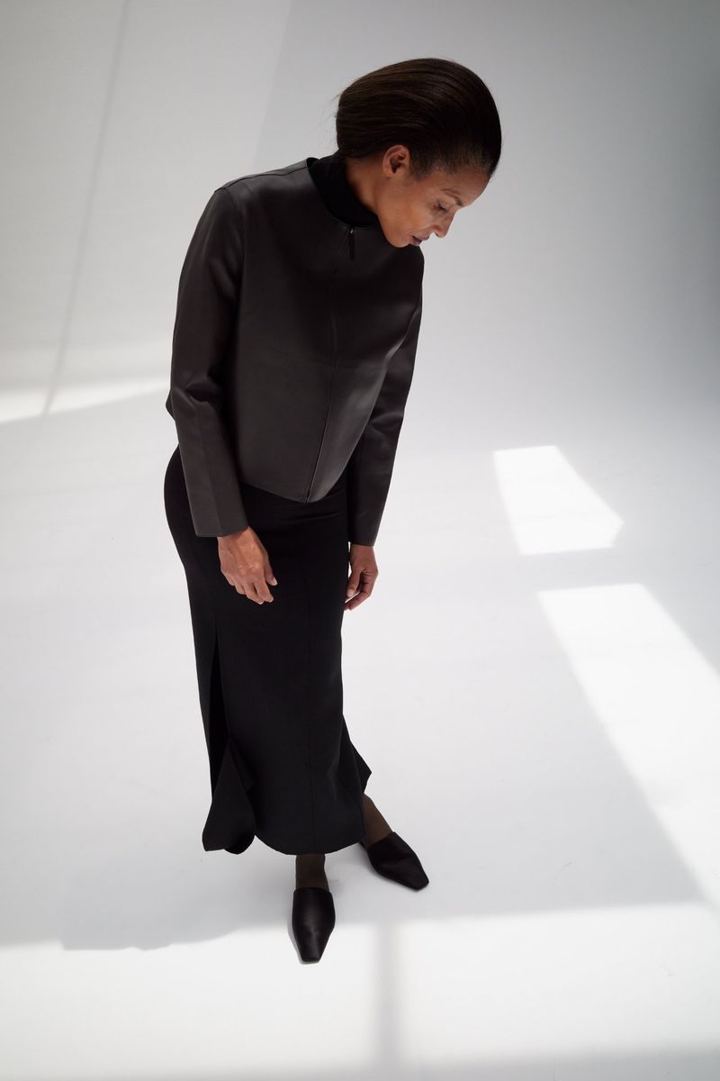 Model Romae Gordon wearing an all-black structured jacket and maxi skirt looking down against a bright white floor with window shadows.