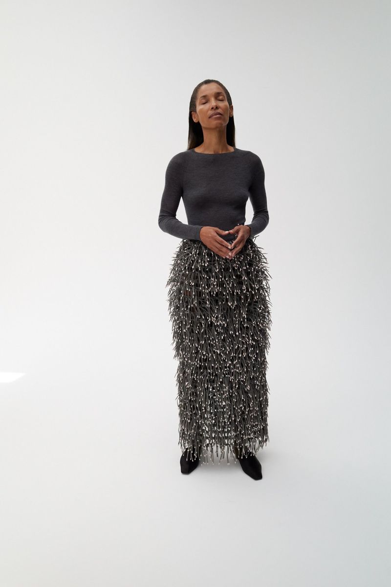 Model Romae Gordon wearing a charcoal long-sleeve knit top and textured fringed maxi skirt against a white studio background.