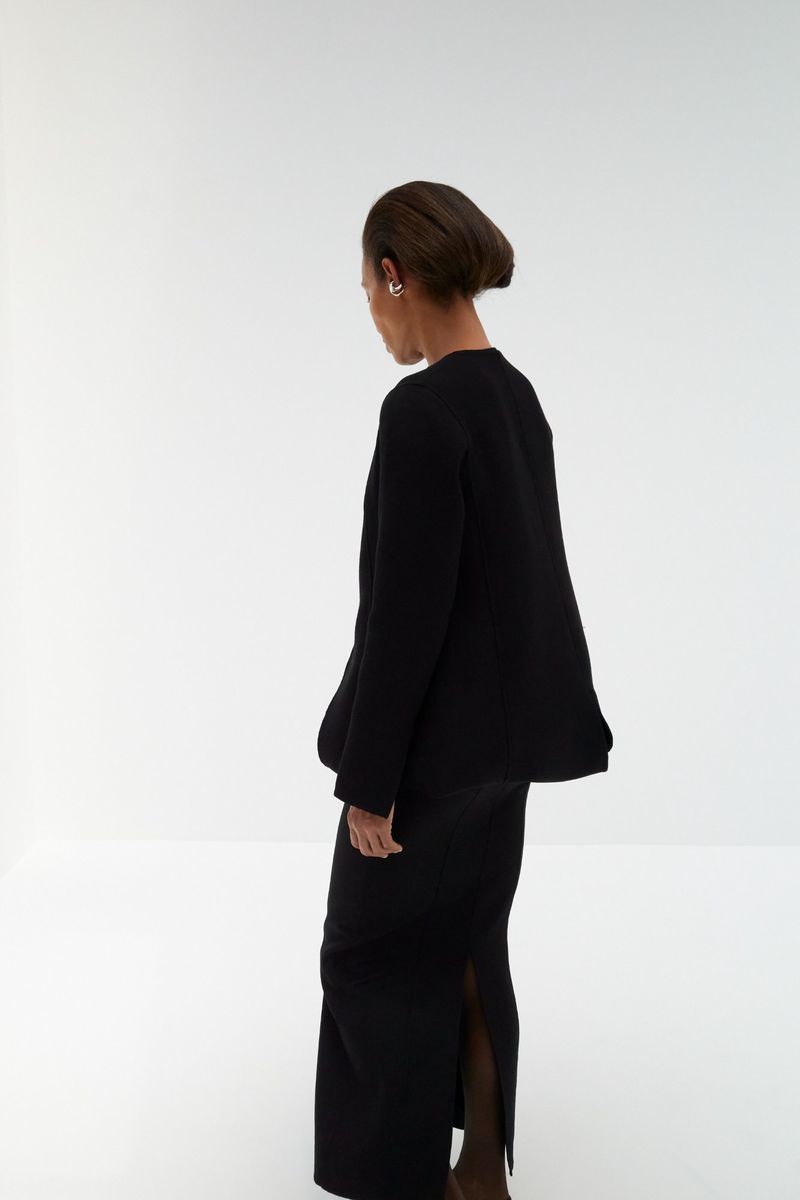 Model Romae Gordon seen from behind wearing a black jacket and matching maxi skirt against a white studio background.