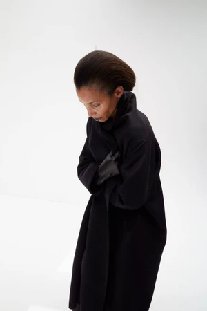 Model Romae Gordon wearing an oversized black coat and gloves, looking downward against a white studio background.
