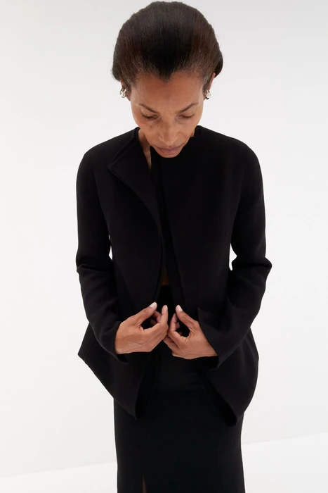 High angle shot of model Romae Gordon in a structured black jacket and skirt against a bright white background.