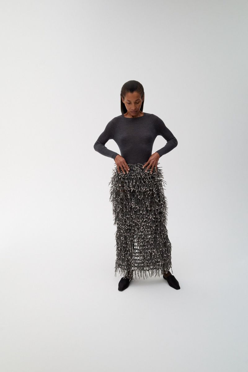 Model Romae Gordon posing in a charcoal long-sleeve top and textured fringe midi skirt against a plain white studio backdrop.