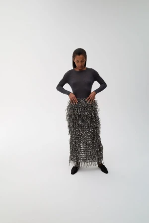 Model Romae Gordon posing in a charcoal long-sleeve top and textured fringe midi skirt against a plain white studio backdrop.