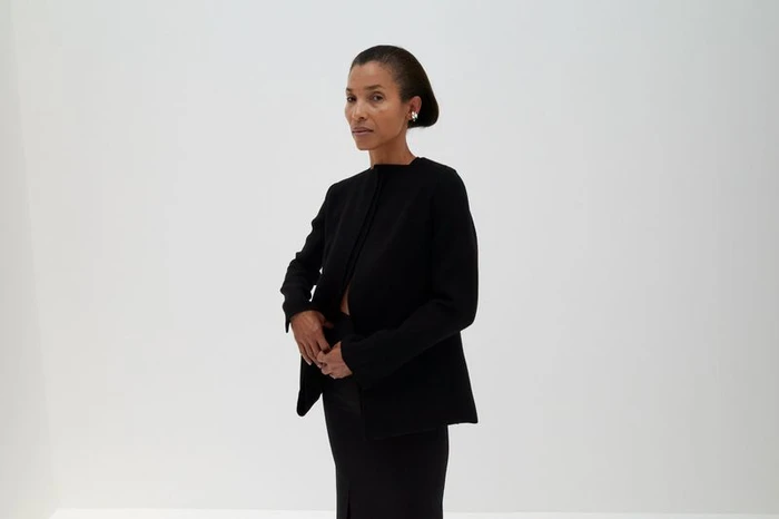 Romae Gordon posing in a structured black collarless jacket and matching skirt against a minimalist white studio backdrop.