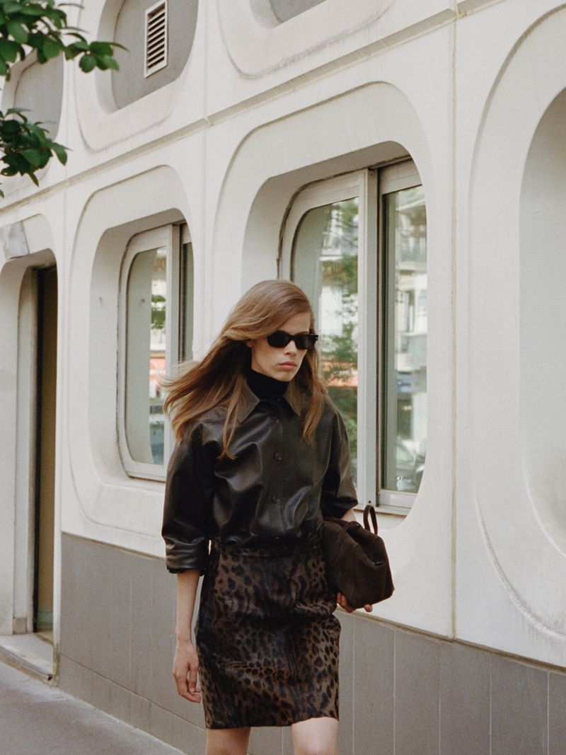 Model wearing a dark leather shirt, leopard print skirt, and sunglasses walking past a white building with rounded windows.
