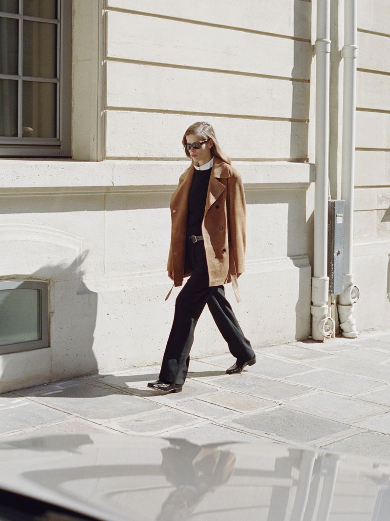 Model Bianca Huisman walking along a sidewalk wearing a camel double-breasted coat, black trousers, and dark sunglasses.