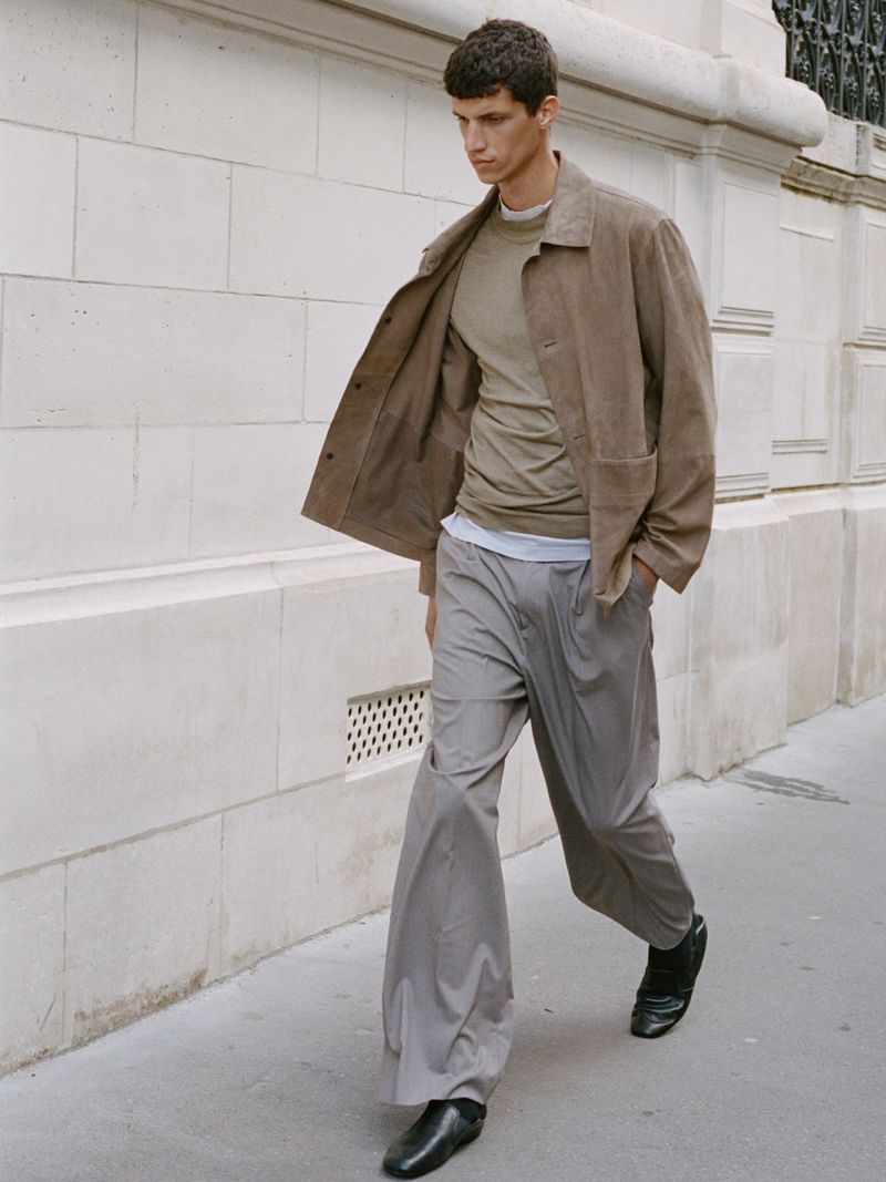 Model walking alongside a stone wall wearing an unbuttoned brown jacket, layered t-shirt, and wide-leg grey trousers.
