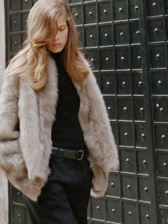 Model Bianca Huisman walking in a taupe fur jacket, black top, and black belt in front of a studded metal gate.