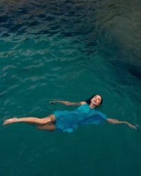 A woman floating peacefully on her back in deep turquoise water wearing a vibrant bright blue dress with arms extended.