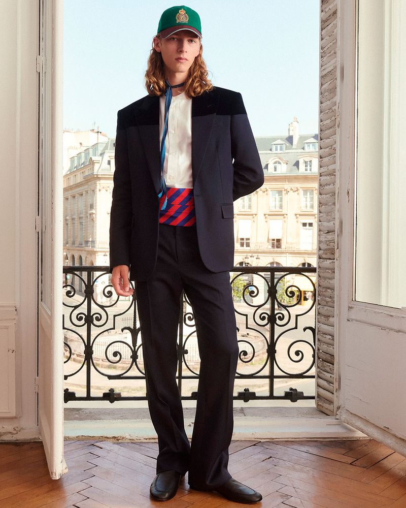 Model wearing a black tailored suit and green baseball cap standing by an open Parisian balcony window.