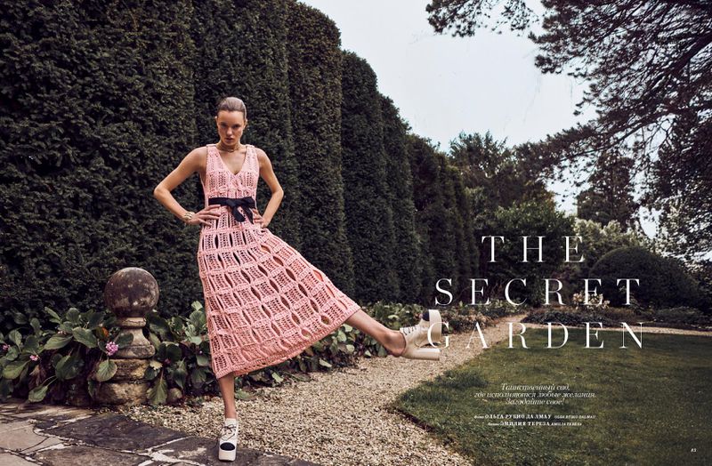 Fashion editorial shot of a female model wearing a pink knit dress, standing in a garden with a tall green hedge backdrop.