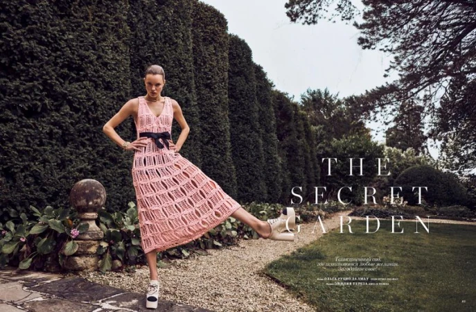 Fashion editorial shot of a female model wearing a pink knit dress, standing in a garden with a tall green hedge backdrop.