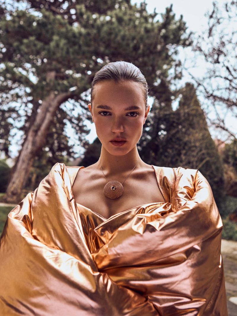 Elegant portrait of a young woman with sleek hair wearing a gold puffy shawl and a round pendant in an outdoor setting with green trees.