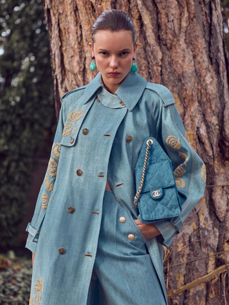 Fashion editorial shot of an adult female model wearing a teal blue coat and pants with a Chanel bag, posing in an outdoor setting.