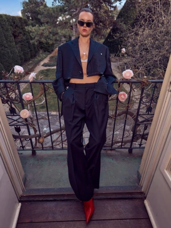 Fashionable woman in cropped suit jacket and wide-leg pants poses on a balcony surrounded by pink roses in an outdoor setting.