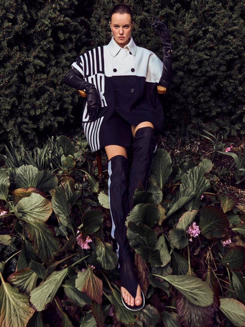 A woman seated in a garden setting wearing a black and white jacket, black gloves and black over-the-knee boots with open-toe heels.