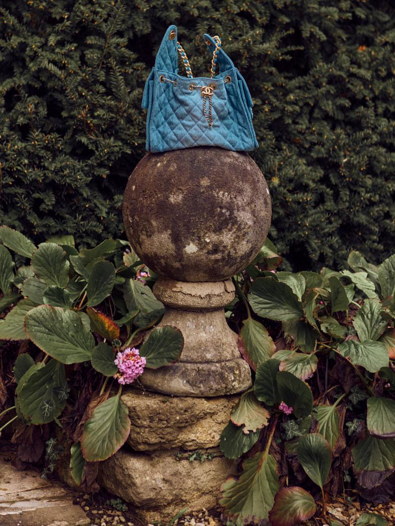 Blue Chanel quilted bucket bag with gold chain straps displayed on a weathered stone pedestal in a lush green garden setting.