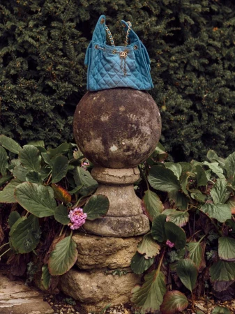 Blue Chanel quilted bucket bag with gold chain straps displayed on a weathered stone pedestal in a lush green garden setting.