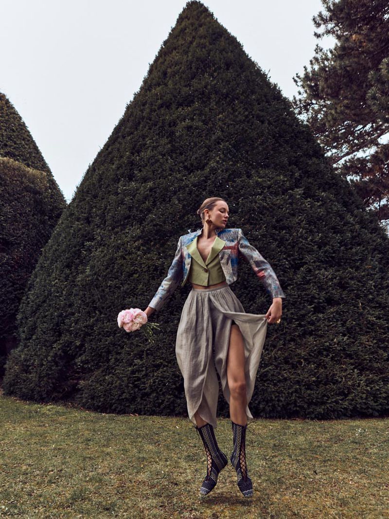 A woman in a jacket, skirt, and boots poses in a garden with green lawns and topiary trees under an overcast sky, holding flowers.