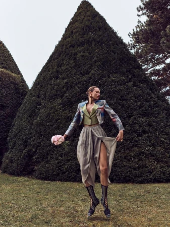 A woman in a jacket, skirt, and boots poses in a garden with green lawns and topiary trees under an overcast sky, holding flowers.