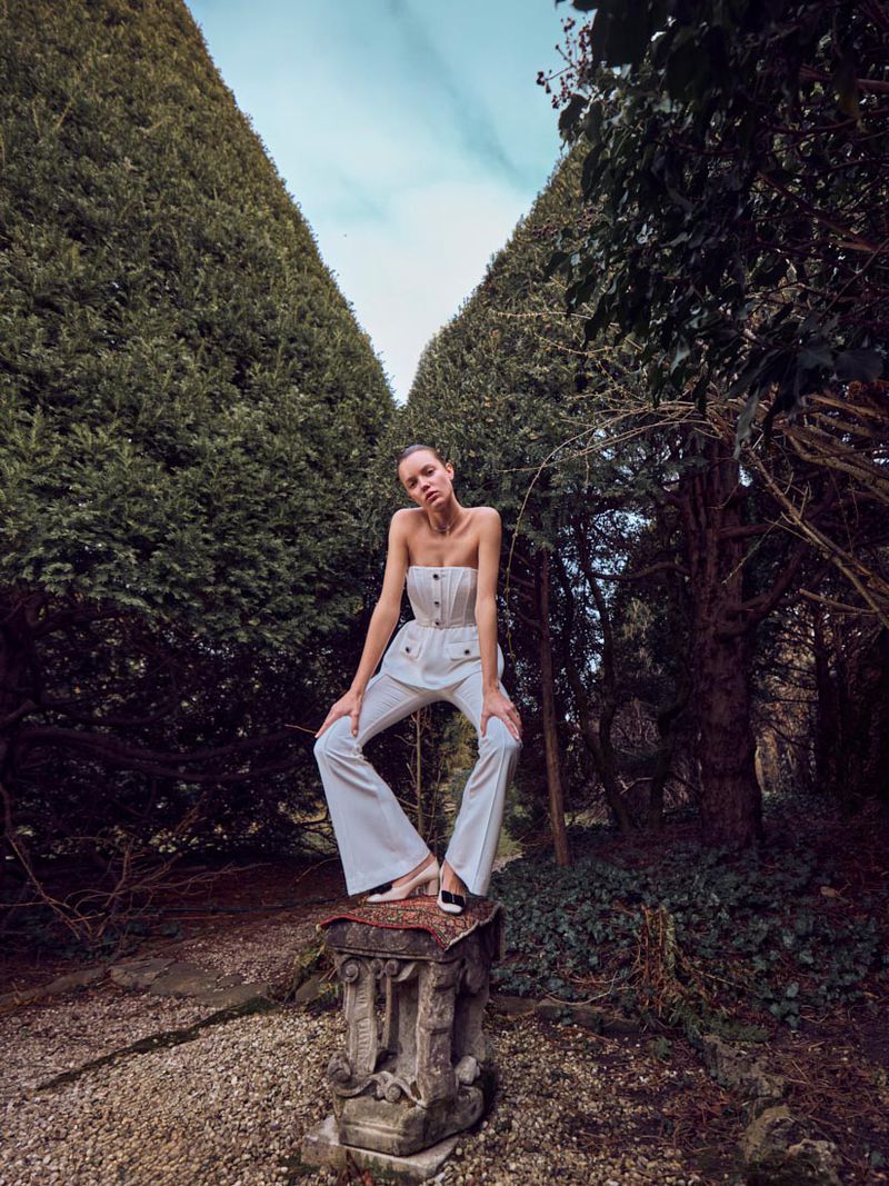Fashion shot of a woman posing on a stone pedestal, wearing a white pantsuit in a garden setting with tall green hedges on a cloudy day.