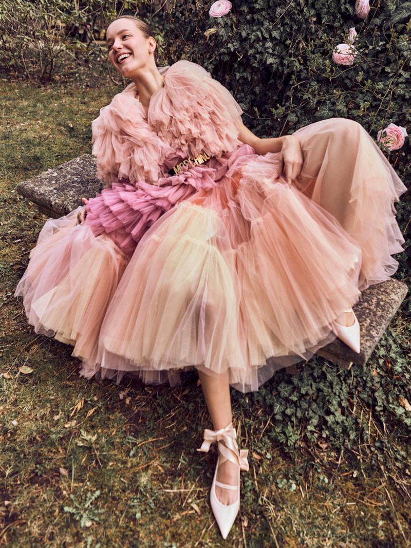 Woman in pink dress poses on stone bench in garden with roses. Elegant fashion portrait in natural light, beauty and style in nature.