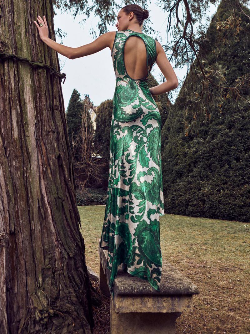 Back view of a woman in a formal green and white gown, standing next to a large tree in a grassy outdoor setting. Fashion photography.
