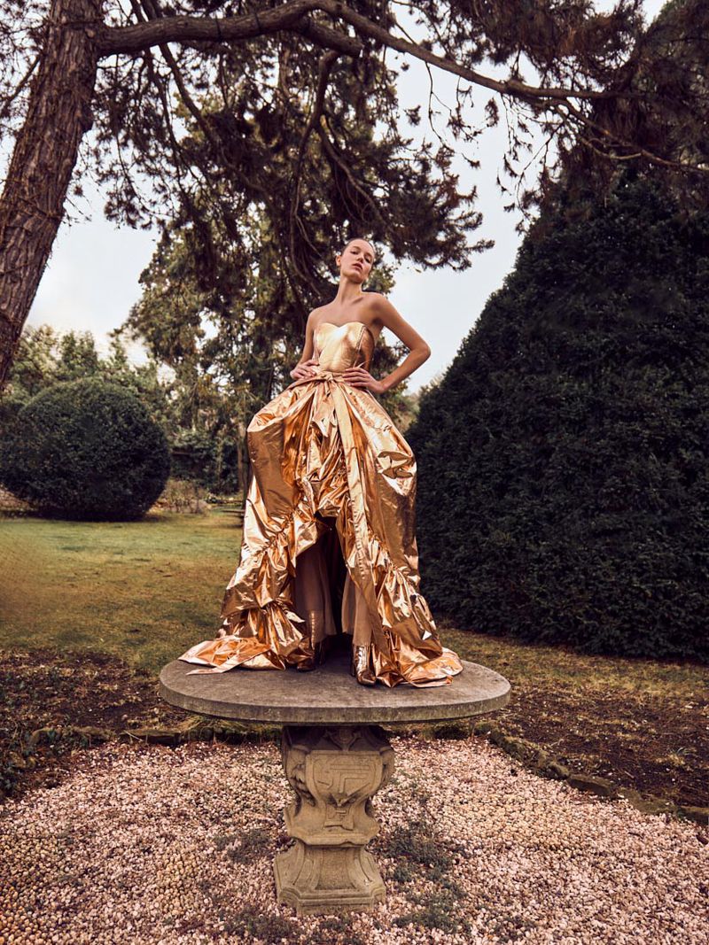 Elegant woman in gold gown posing on stone pedestal in outdoor garden with trees and manicured bushes, fashion editorial shot.