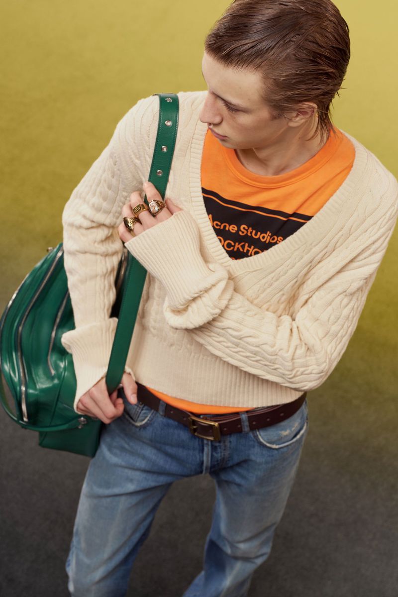 High-angle view of a man wearing a cream cable-knit sweater over an orange graphic t-shirt and blue jeans with a green bag.