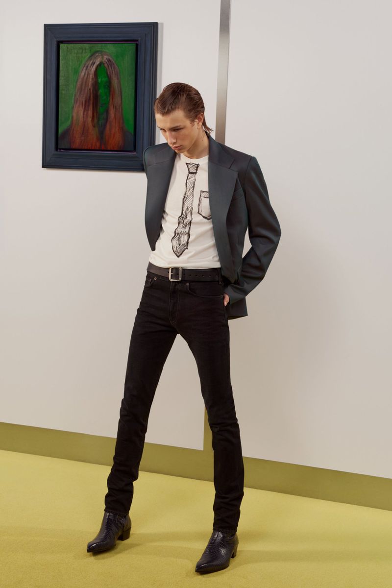 Model wearing a dark blazer, graphic t-shirt, and black jeans standing in a gallery interior with a painting on the wall.