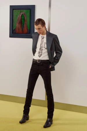 Model wearing a dark blazer, graphic t-shirt, and black jeans standing in a gallery interior with a painting on the wall.