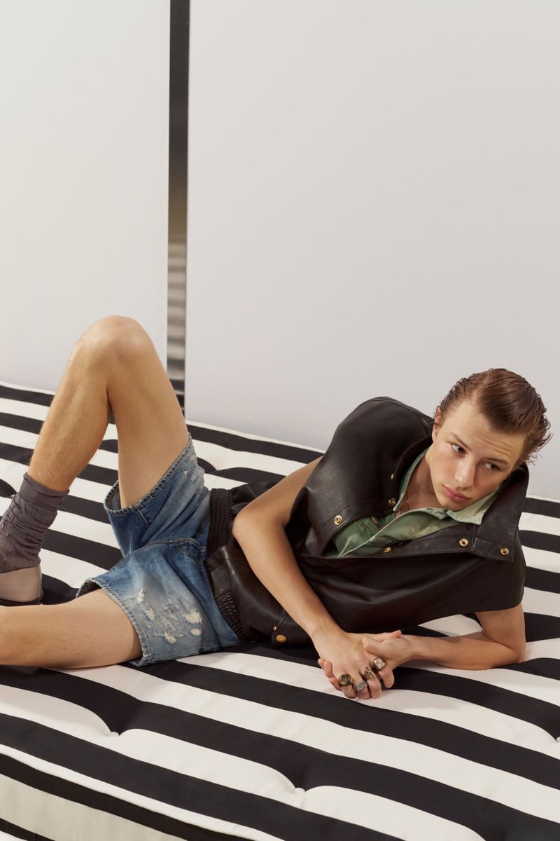 Model Pavlo Stinyk reclining on a black-and-white striped daybed wearing a black leather vest and denim shorts.